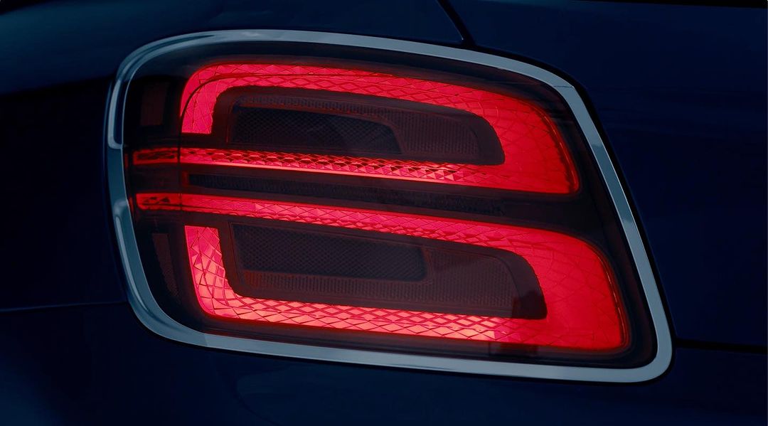 Close-up of a modern car taillight with a geometric design, featuring bright red light bars and a silver trim, set against a dark background.