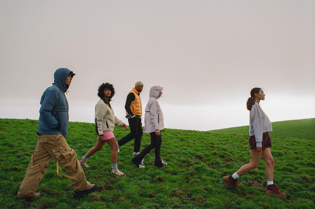 Five people walk through a grassy field under an overcast sky. They are wearing outdoor clothing including jackets and vests.