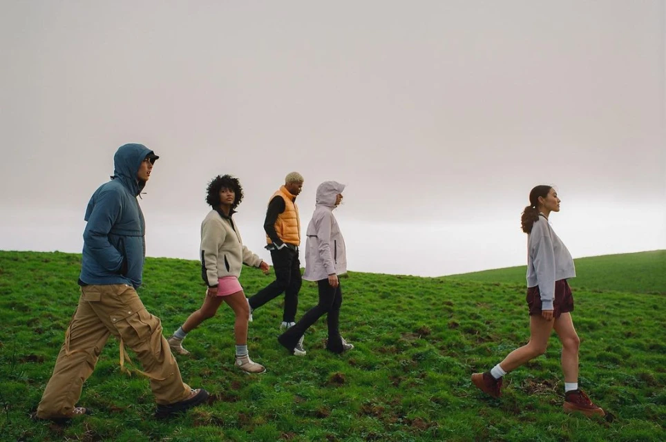 Five people walk through a grassy field under an overcast sky. They are wearing outdoor clothing including jackets and vests.