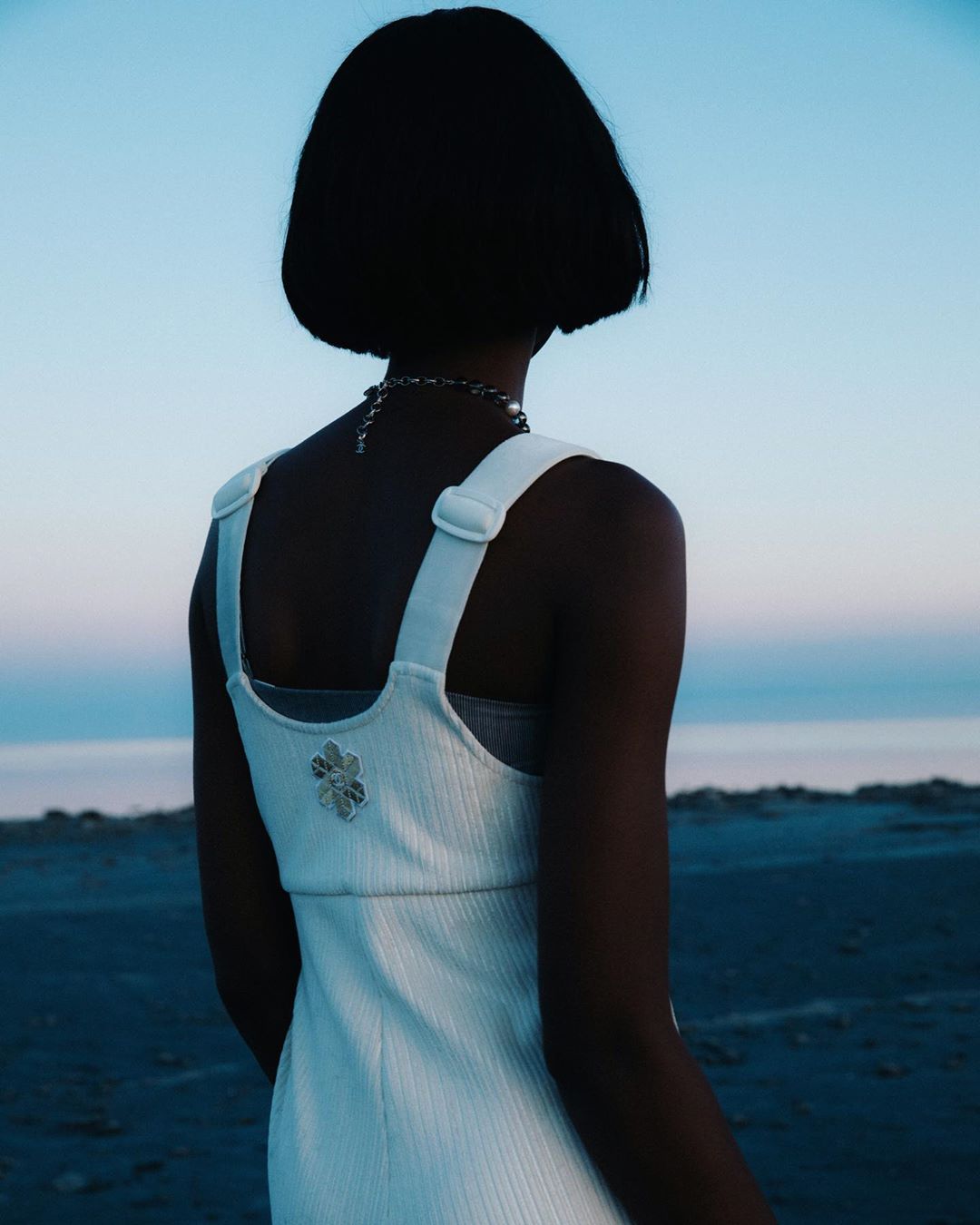 Back view of a slender adult woman with a bob haircut in a light-colored dress and jewelry outdoors under a twilight sky.