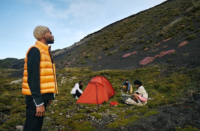 A group of people camping on a grassy hillside with a mountain in the background; one man in an orange vest stands in the foreground.