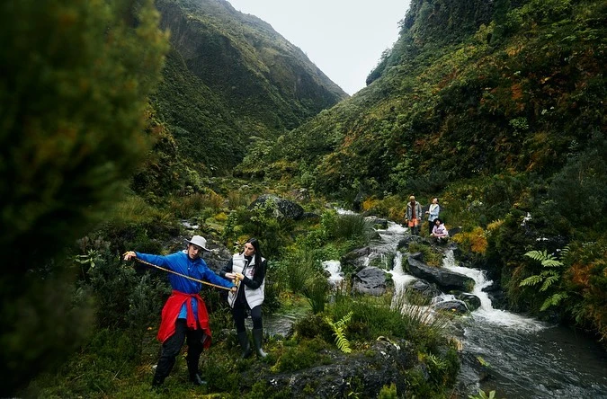 Group of people in a lush green mountain valley with a stream; man and woman hold a rope in the foreground, mountains visible.