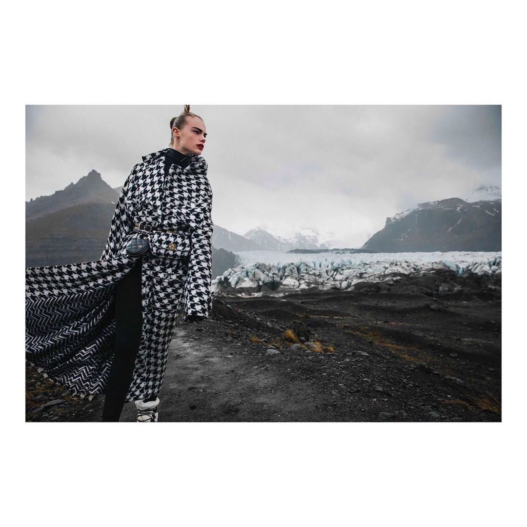 Woman in houndstooth coat stands on rocky terrain with glacier and mountains in the background. Fashion editorial outdoor shot.