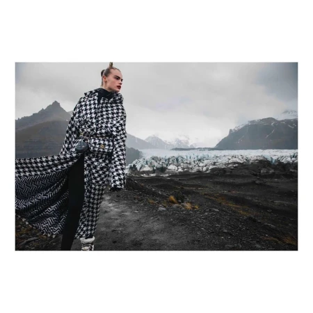 Woman in houndstooth coat stands on rocky terrain with glacier and mountains in the background. Fashion editorial outdoor shot.
