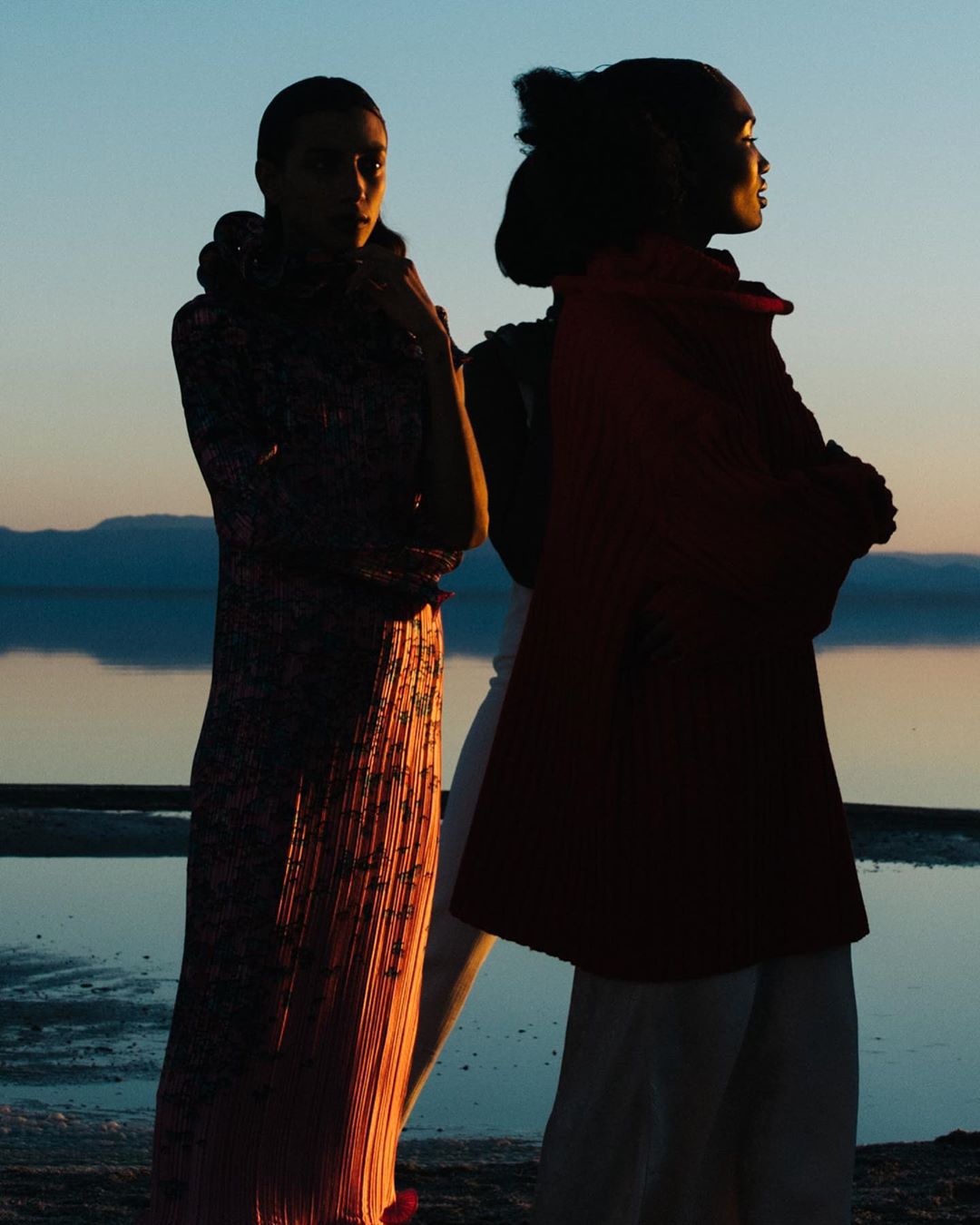 Two fashion models stand on a beach at sunset. Silhouettes are set against the sky and water. The models are wearing long dresses.