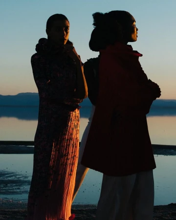 Two fashion models stand on a beach at sunset. Silhouettes are set against the sky and water. The models are wearing long dresses.