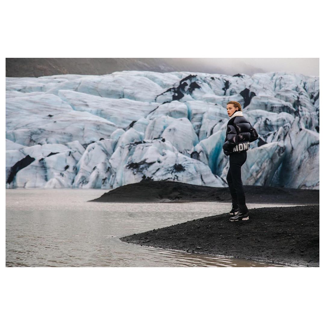 Woman in a puffer jacket standing near the water's edge with a view of a glacier, creating a dramatic landscape photograph.