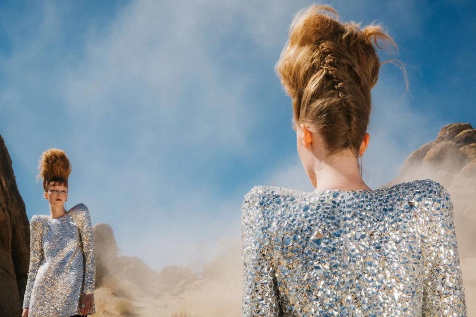 Two fair-skinned adult women with elaborate hairstyles and silver sequin dresses in a desert setting under a blue sky. Fashion shot, full view.