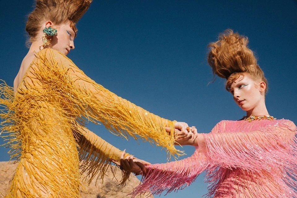 Two women with red hair pose in pink and yellow fringe dresses outdoors against a blue sky, holding hands in a fashion editorial.