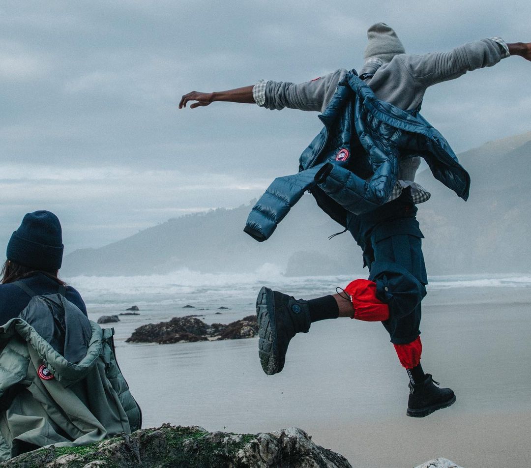 Person in mid-air jump on a beach with a cloudy sky, wearing a blue Canada Goose jacket and dark clothing with red leg coverings.