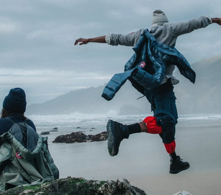 Person in mid-air jump on a beach with a cloudy sky, wearing a blue Canada Goose jacket and dark clothing with red leg coverings.