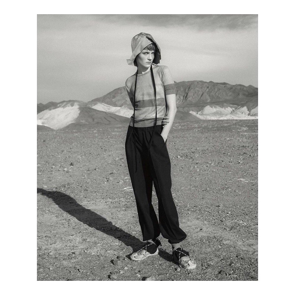 Monochrome fashion portrait of a model wearing a bucket hat, striped short sleeve top, and dark trousers in a desert landscape