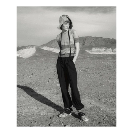 Monochrome fashion portrait of a model wearing a bucket hat, striped short sleeve top, and dark trousers in a desert landscape