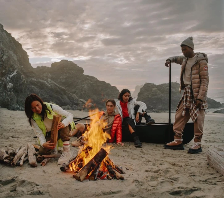 Four people sit on a beach near a fire, wearing jackets and warm clothes. There is driftwood on the beach, a cloudy sky, and ocean.