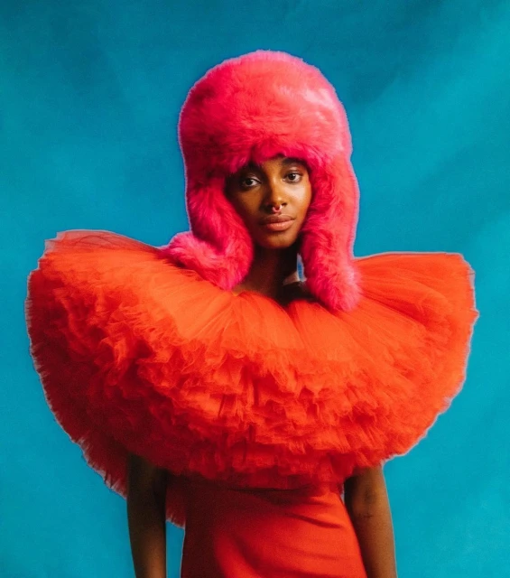 Woman wearing a red dress and fluffy pink headpiece in a studio portrait with a blue background for a fashion editorial.