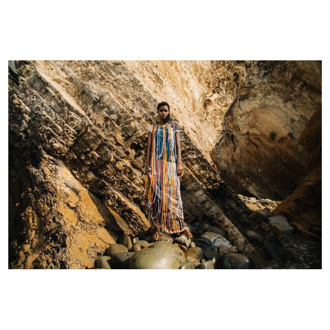 Fashion shot of a Black woman in a patterned dress with draped cords, standing on rocks in front of a cliff face with layered rock strata.
