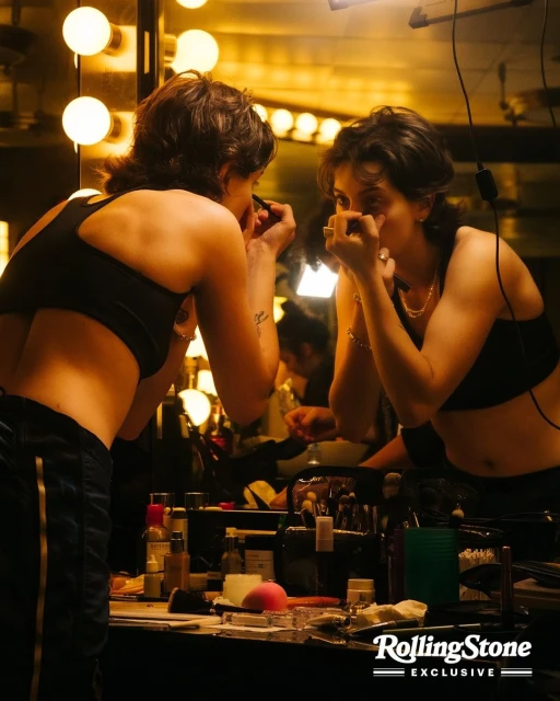 Woman applying makeup while looking in mirror in dressing room with vanity; makeup brushes, skincare products on table.