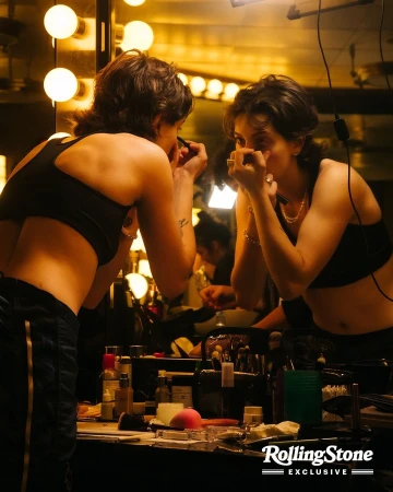Woman applying makeup while looking in mirror in dressing room with vanity; makeup brushes, skincare products on table.