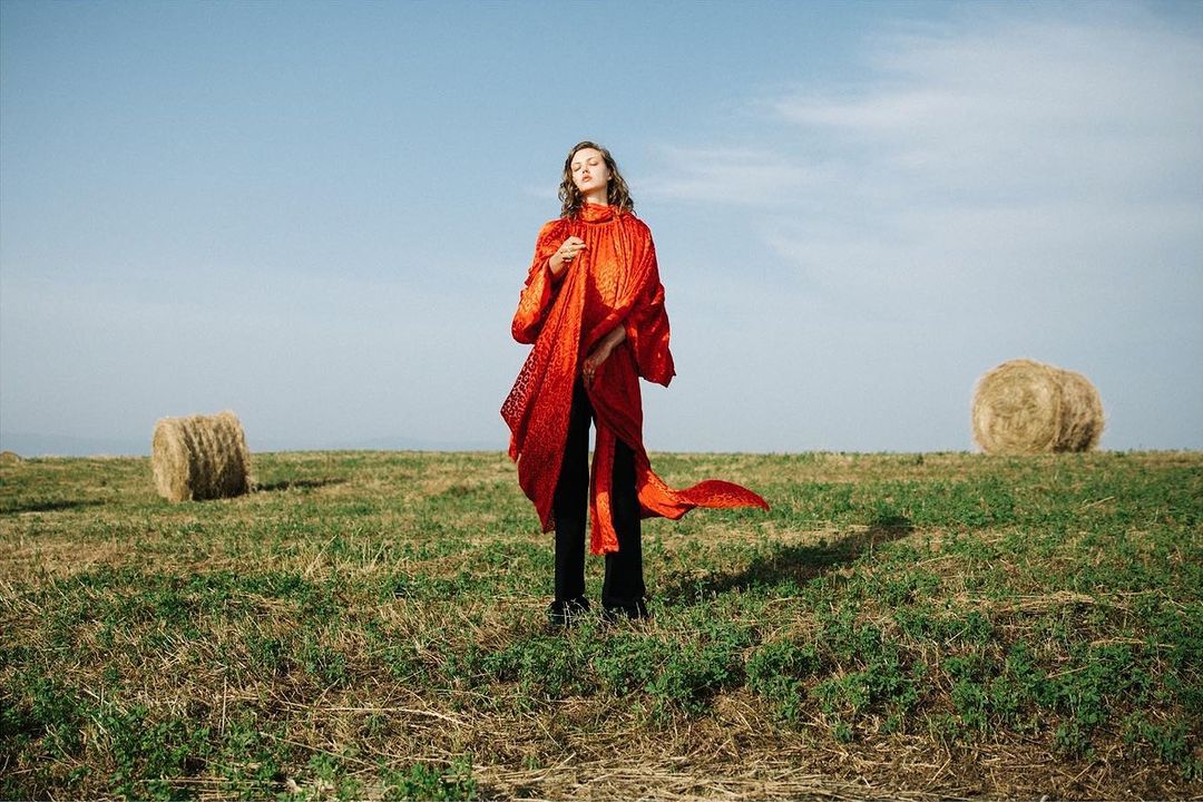 A woman in a red coat stands in a grassy field near hay bales beneath a clear sky, fashionably posed in an outdoor setting.