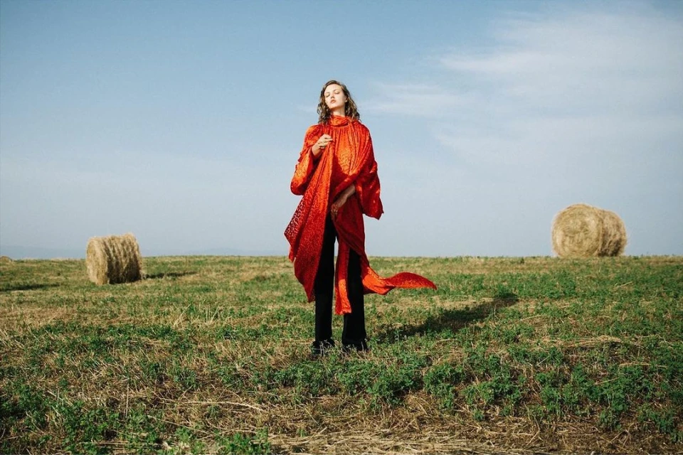 A woman in a red coat stands in a grassy field near hay bales beneath a clear sky, fashionably posed in an outdoor setting.