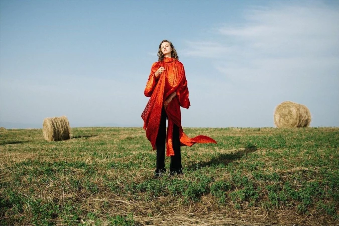 A woman in a red coat stands in a grassy field near hay bales beneath a clear sky, fashionably posed in an outdoor setting.
