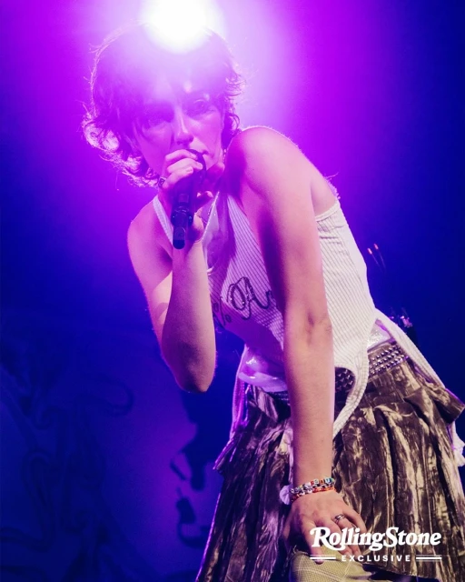 A young adult female singer performs on stage with a microphone in hand, wearing a white top and long skirt, under bright purple stage lights.