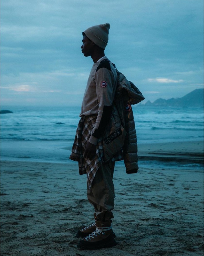 Profile shot of an African-American man in outdoor apparel standing on a beach in front of the ocean, wearing a beanie and a jacket.