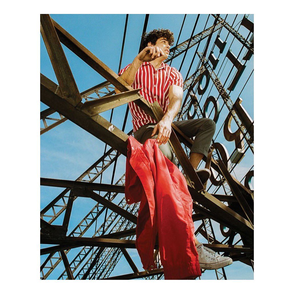 Young man in striped shirt sitting on steel structure with red jacket, against blue sky. Urban setting with strong lines.