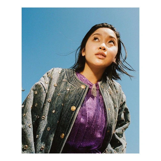 Portrait of a young Asian woman with dark hair wearing a denim jacket and purple shirt, looking up against a bright blue sky.