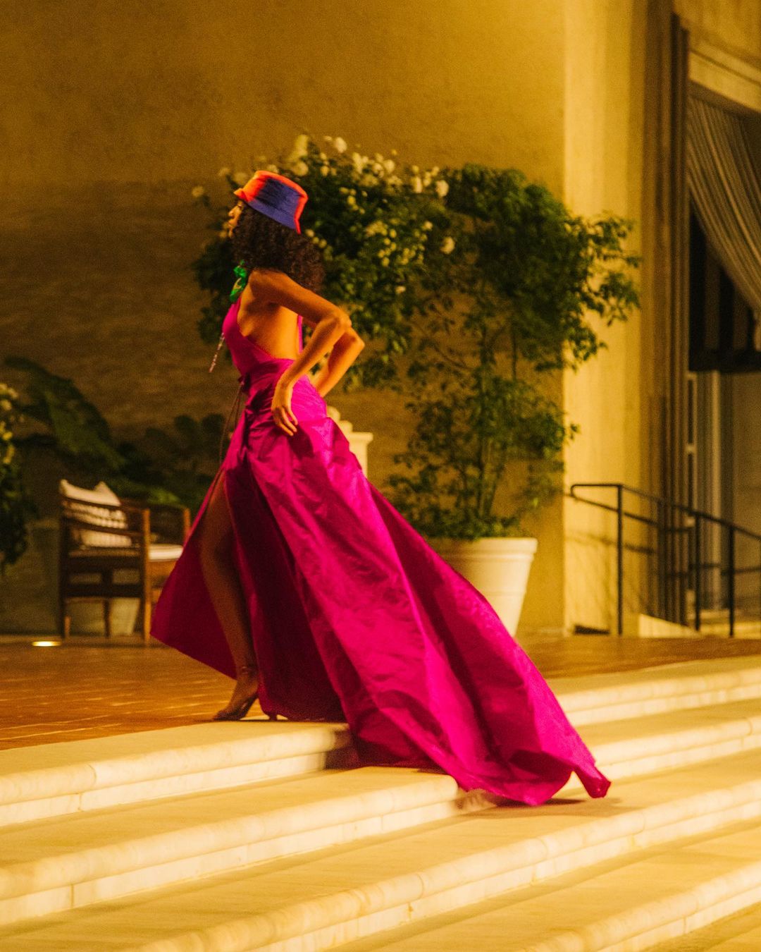 Woman in a magenta gown and colorful hat walks down a set of stairs surrounded by white flowers in an outdoor setting.