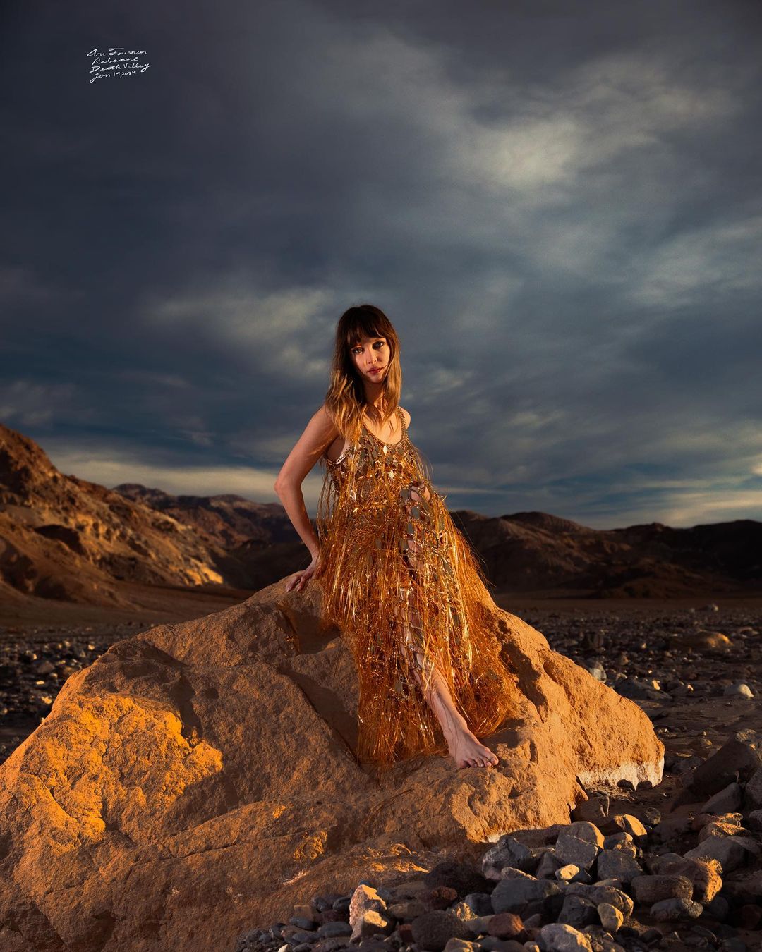 A woman in a shimmering gold dress sits on a rock in a desert landscape with mountains and a cloudy sky providing the backdrop.