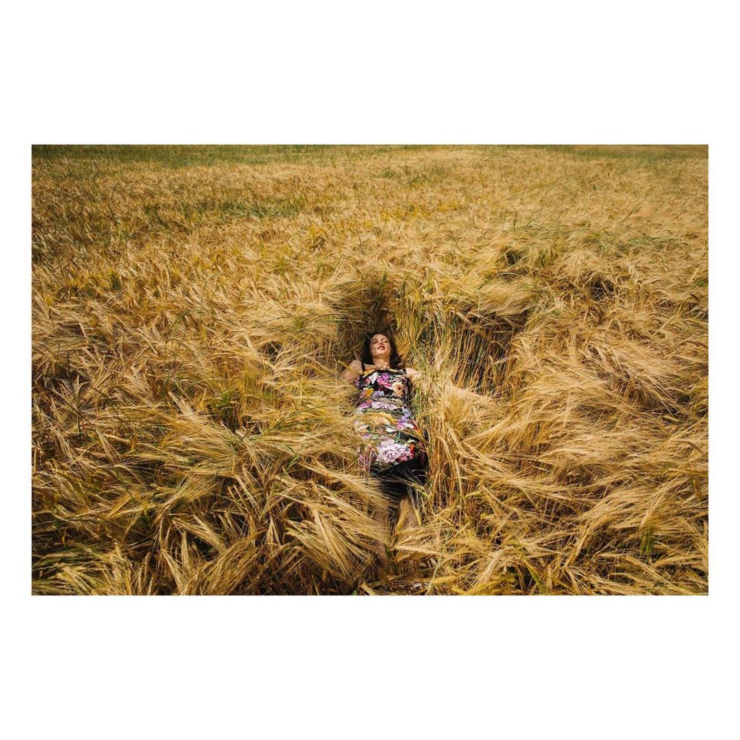 Woman in floral dress lies in a sunlit wheat field, captured in a wide shot showing golden hues and a peaceful rural scene.