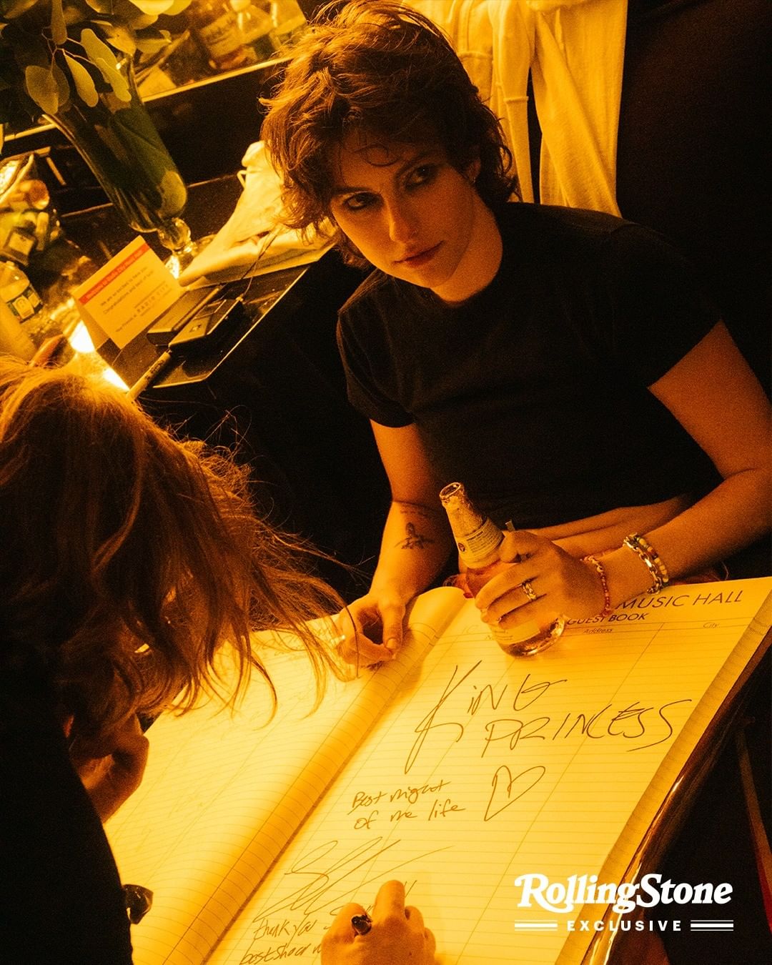 A young adult woman with short brown hair in a black shirt holds a beer while signing a large guest book next to another person indoors.