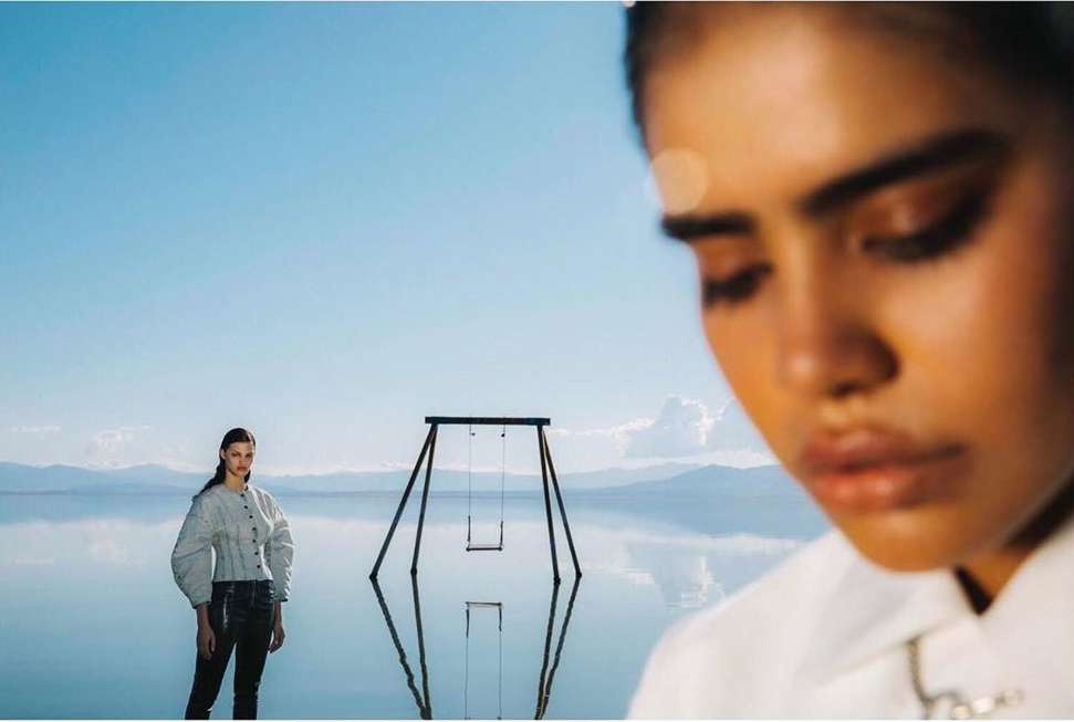 Fashion shot of two women standing in shallow water under a blue sky with a swing set reflected in the water and mountains in the distance.