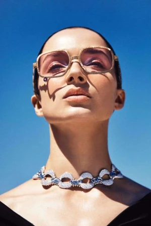 Eye-level portrait of a woman wearing gold-rimmed sunglasses and a diamond necklace against a clear, light blue sky backdrop.