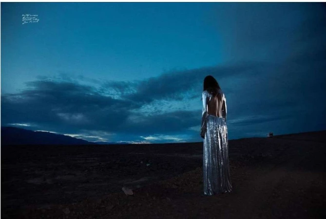 An adult female with long hair stands in the desert wearing a shimmering, backless gown under a dramatic dark blue, cloudy sky.