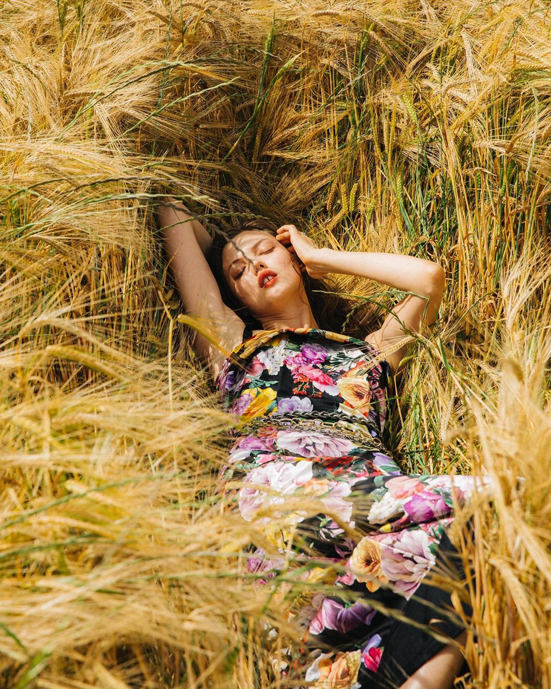 Young woman in a floral dress lies in a field of golden wheat, her hands near her head, captured in natural light.