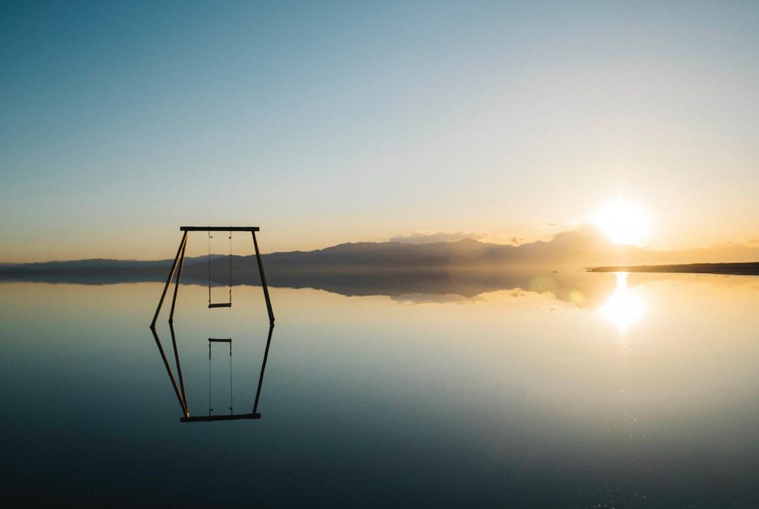 Swing set sits on a body of water mirroring the sky and the horizon, with soft sunlight creating a dreamlike, serene atmosphere.