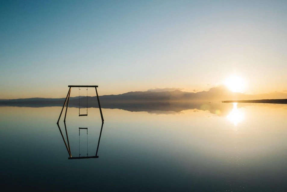 Swing set sits on a body of water mirroring the sky and the horizon, with soft sunlight creating a dreamlike, serene atmosphere.