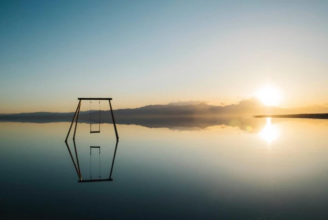 Swing set sits on a body of water mirroring the sky and the horizon, with soft sunlight creating a dreamlike, serene atmosphere.