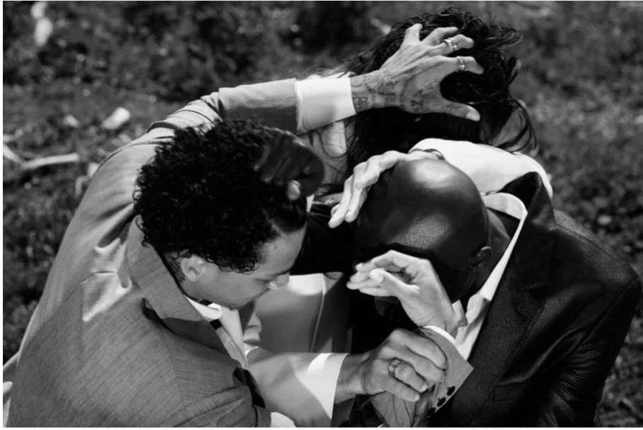 Three people in suits embrace closely in a black-and-white photograph taken outdoors, focusing on the intimate human connection and formal attire.