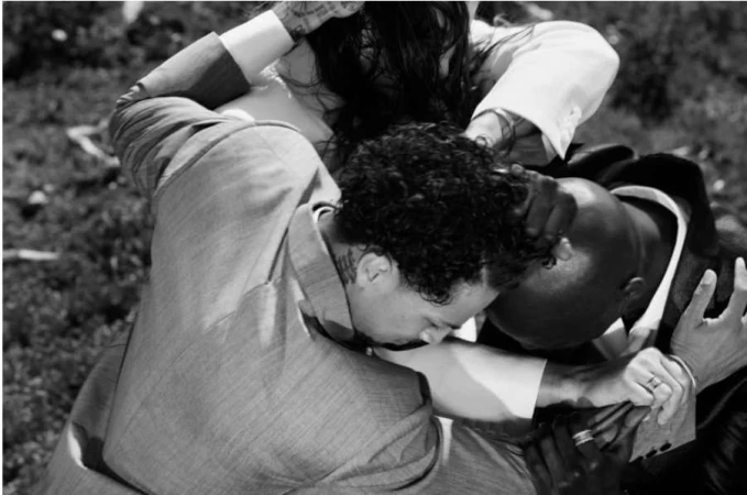 Three people embrace, showcasing connection and support, wearing suits, outdoors in natural light, with a shallow depth of field in monochrome.