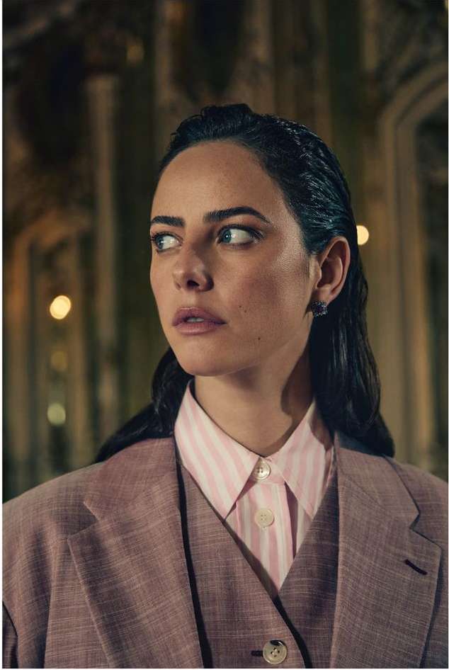 Portrait of fair-skinned adult female in a pink suit and striped shirt, looking off-camera in a dimly lit interior with arched openings.