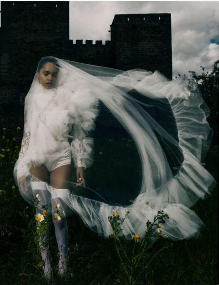 A female fashion model poses in a romantic white gown and veil outdoors near a castle, with flowers in the foreground and cloudy sky.