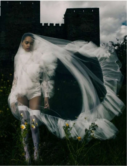 A female fashion model poses in a romantic white gown and veil outdoors near a castle, with flowers in the foreground and cloudy sky.