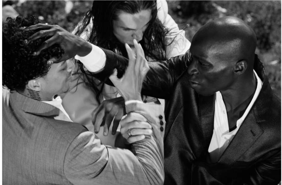 Black and white portrait of three adult men in suits, touching each other's heads and arms in an outdoor setting.