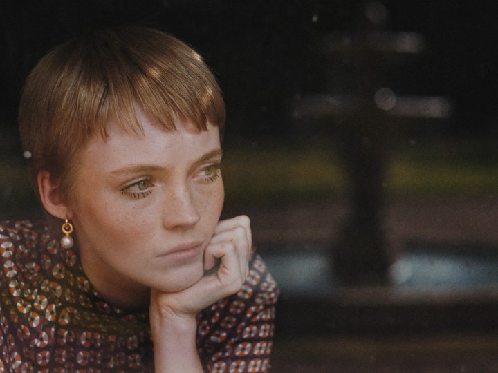 Close-up portrait of a young woman with short, light brown hair wearing a patterned shirt and pearl earrings, gazing pensively in soft lighting.