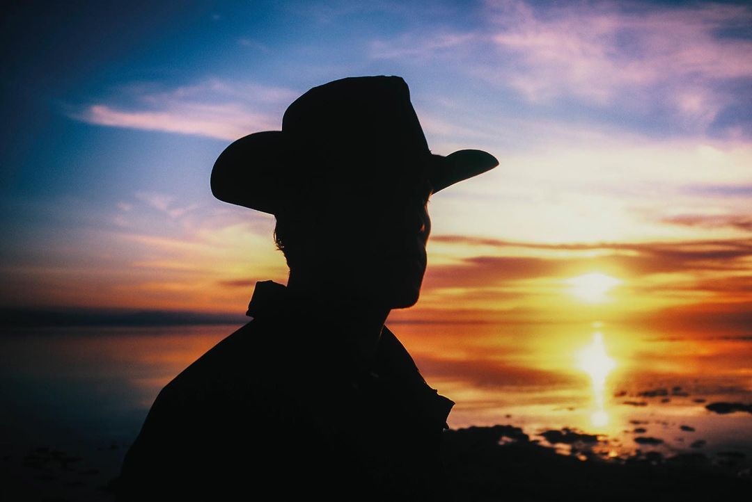 Silhouette of a person wearing a cowboy hat looking out at a body of water during a vibrant sunset with pink and orange hues.