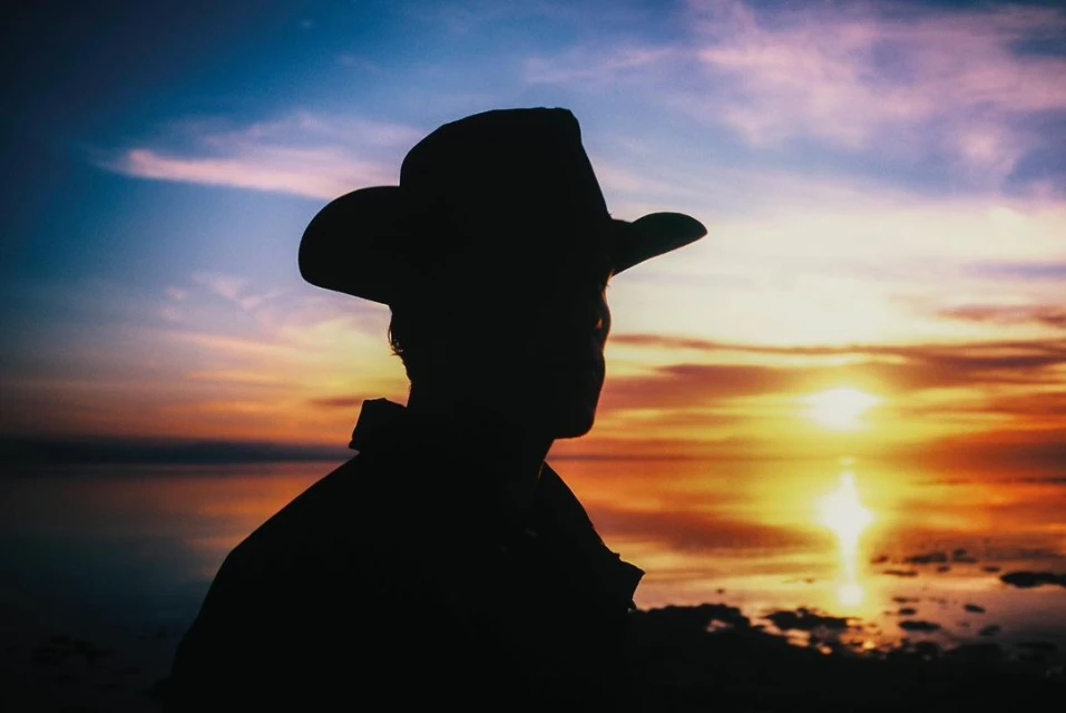 Silhouette of a person wearing a cowboy hat looking out at a body of water during a vibrant sunset with pink and orange hues.