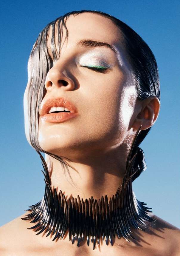 Close-up beauty shot of a woman with wet hair, closed eyes, and decorative neckpiece against a clear blue sky background.
